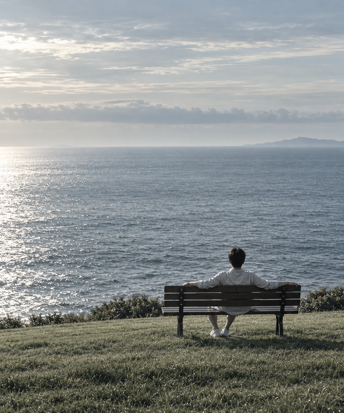 A person seen from behind, seated on a bench facing the sea.